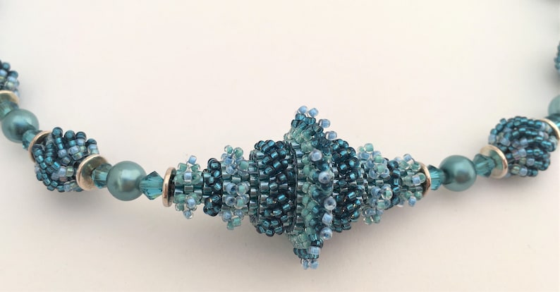 Pu&ograve; includere: Una collana di perline blu e verdi con un grande perno a spirale al centro.