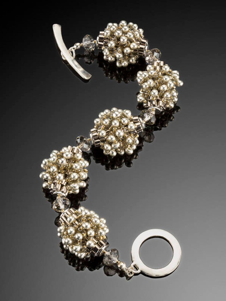 May include: A beaded bracelet with a silver toggle clasp. The bracelet features clusters of small, round, gold-coloured beads interspersed with clear crystal beads. The bracelet is displayed on a reflective black surface.