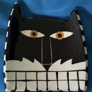 May include: A square ceramic dish featuring a stylized black cat face with large orange eyes, a white muzzle, and a wide grin with visible teeth. The sides of the dish are decorated with a black and white checkered pattern on a glossy surface.