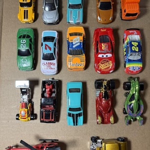 May include: A collection of seventeen die-cast toy vehicles arranged in rows, including various race cars, a bright orange truck, a red helicopter, and several sports cars in yellow, silver, blue, red, green, and gold colors.