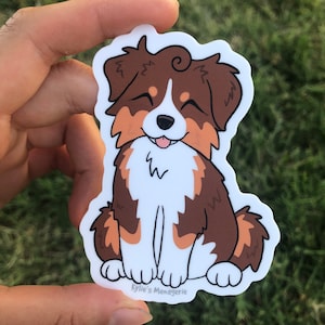 May include: A sticker featuring a cartoon Australian Shepherd dog with brown, white, and orange fur. The dog is sitting with its tongue sticking out and its eyes closed. The sticker has the text "Kylie's Menagerie" at the bottom.