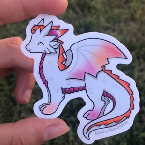 Puede incluir: Una pegatina de dragón blanco con detalles en rosa y naranja. El dragón tiene una cola rosa y blanca, alas rosa y blancas y una melena rosa y naranja. La pegatina tiene el texto "Kylie's Menagerie" en la parte inferior.