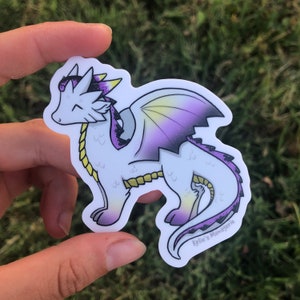 May include: A white dragon sticker with purple, yellow, and black accents. The dragon has a friendly expression, with a yellow chain around its neck and purple wings. The sticker is held by a hand against a green background. The text "Kylie's Menagerie" is visible.