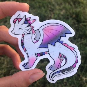 May include: A white dragon with pink, blue, and purple scales and a galaxy-like pattern on its wings. The dragon is standing on all fours with its wings spread out. The dragon has a pink and blue crown on its head. The sticker has the text "Kylie's Menagerie" at the bottom.