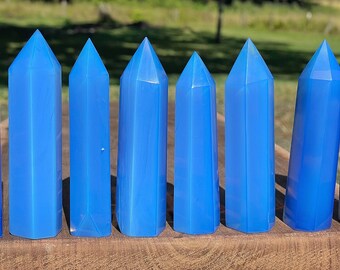 May include: Eight blue crystal points, also known as wands, are arranged in a row on a wooden surface. The points are all the same shape and size, and they have a smooth, polished finish.