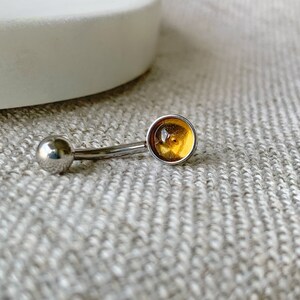以下が含まれることがあります： 丸い琥珀色の宝石が付いたシルバーのボディピアス。宝石の中に金色の斑点がある。