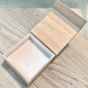 以下が含まれることがあります： 木目調の内側にベージュの布で覆われた箱。箱が開いており、中央に小さな窪みのある白いベルベットの裏地が付いている。