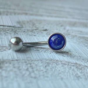 May include: A silver body piercing with a blue gemstone. The gemstone is round and has a silver setting. The piercing is curved and has a ball on one end.