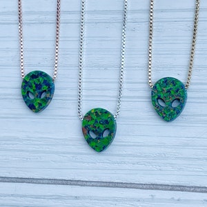 May include: Three silver chains with green and blue alien head pendants. The chains are on a white wooden background.