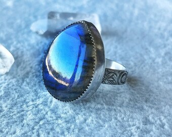 May include: A silver ring with a blue labradorite gemstone set in a bezel setting. The band is decorated with an intricate floral design.