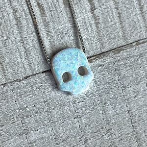 May include: A silver chain necklace with a white opal skull pendant. The skull is carved with intricate details and has a blue iridescent sheen.