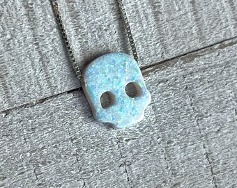 May include: A silver chain necklace with a white opal skull pendant. The skull is carved with intricate details and has a blue iridescent sheen.