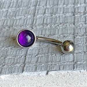 May include: A silver belly button ring with a purple gem. The ring is curved and has a small ball on the end.