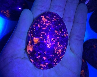 May include: A close-up of a purple and pink gemstone with a rough, textured surface. The stone is illuminated with a UV light, highlighting the pink and orange inclusions within the stone.