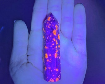 May include: A purple and orange crystal point with a rough, textured surface. The crystal is illuminated with a UV light, highlighting the orange inclusions.
