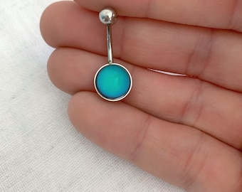 以下が含まれることがあります： 丸い青い宝石が付いたシルバーのへそピアス。宝石はわずかに虹色に輝いています。