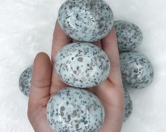 May include: A collection of polished, oval-shaped stones. The stones have a light blue base color with speckled gray and black patterns. The stones are smooth and appear to be natural, possibly gemstones or decorative rocks.