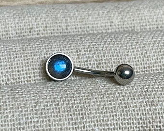 May include: A curved, silver-colored belly button ring. One end features a round, bezel-set, blue-gray gemstone. The other end has a small, silver-colored ball. The jewelry is displayed on a textured, light gray surface.