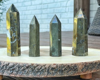 May include: Four green and gold pyrite crystal points on a wooden slice. The points are arranged in a row, with the largest point on the left and the smallest point on the right.