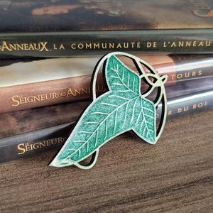 May include: A green and white leaf-shaped pin with a detailed leaf design. The pin is set against a backdrop of movie cases with French text, including "SEIGNEUR DES ANNEAUX". The pin is approximately 2 inches in length.