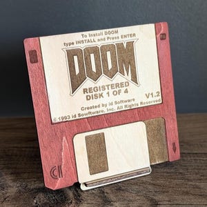 Peut inclure: Un lecteur de disquette en bois avec le logo DOOM et les détails du jeu. Le disque est rouge avec une étiquette centrale de couleur claire qui indique "DOOM REGISTERED DISK 1 OF 4". Le texte "To Install DOOM type INSTALL and Press ENTER" est également visible.