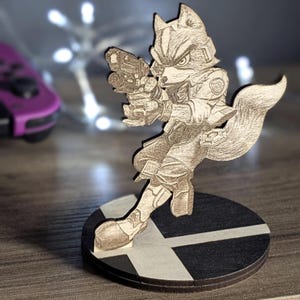 Peut inclure: Figurine en bois de Fox McCloud de Star Fox, tenant un blaster, sur une base circulaire noire et blanche. La figurine est détaillée avec des lignes découpées au laser, montrant l'uniforme et les traits du personnage. L'arrière-plan est flou.