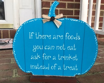 May include: A blue wooden pumpkin-shaped sign with white text that reads, "If there are foods you can not eat ask for a trinket instead of a treat." The sign has a brown base and a raffia bow.