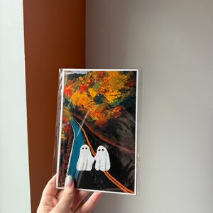 May include: A greeting card featuring an autumn landscape with a road and two white ghost figures. The artwork has a vibrant color palette of orange, yellow, blue, and black. The card is held in a clear plastic sleeve.