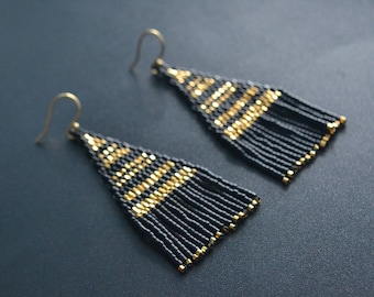 Pode incluir: Um par de brincos longos de contas pretas e douradas. Os brincos têm forma triangular e têm uma franja de contas pretas.