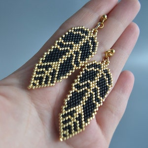 Könnte beinhalten: Ein Paar Hängeohrringe mit einem schwarzen und goldenen Blattdesign. Die Ohrringe bestehen aus kleinen Perlen und haben einen goldfarbenen Metallhaken.
