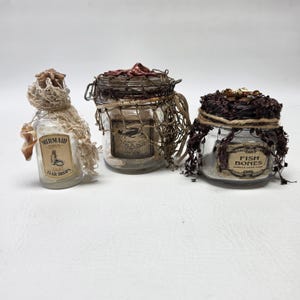 May include: Three glass jars with cork or metal lids, decorated with netting, twine, and shells. The labels read Mermaid Tear Drops, Jaws of Crabs, and Fish Bones. Each jar contains small items and features a distinct nautical-themed embellishment.