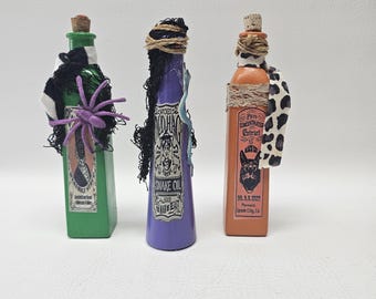May include: Three decorative potion bottles in green, purple, and orange. Each bottle has a cork stopper and is adorned with fabric, twine, and a spider. The bottles feature labels with text and illustrations, evoking a Halloween theme.