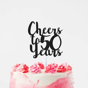 May include: Black glitter cake topper with the text "Cheers to 50 Years" on a pink and white frosted cake.