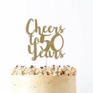 May include: Gold glitter cake topper with the text "Cheers to 50 Years" on a white cake with brown frosting.