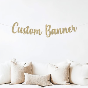 May include: A gold glitter "Custom Banner" hangs above a white sofa with beige pillows. The banner is strung on a thin white string. The sofa sits on a neutral-colored rug. A vase with tall grass is on the left.