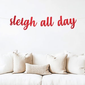 May include: Red glitter banner with the text "sleigh all day" hanging above a white couch with beige pillows.
