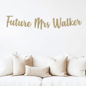 May include: Gold glitter banner with the text "Future Mrs. Walker" hanging on a white wall above a white couch with beige pillows.