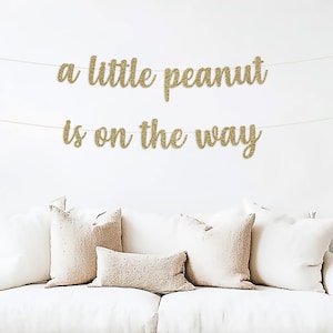 Puede incluir: Una pancarta de brillo dorado con el texto "a little peanut is on the way" colgada en una pared blanca sobre un sof&aacute; blanco con almohadas.
