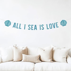 May include: A banner with the phrase "ALL I SEA IS LOVE" in turquoise glitter letters, flanked by seashell designs. The banner is strung above a white sofa with beige pillows, creating a coastal theme.