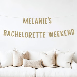 May include: Gold glitter banner with the text "Melanie's Bachelorette Weekend" hanging on a white wall above a white couch with beige pillows.