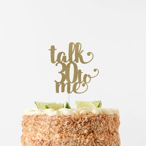 May include: A white cake decorated with shredded coconut and lime slices, topped with a gold glitter cake topper that reads "talk 30 to me".