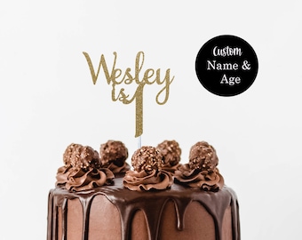 May include: Gold glitter cake topper with the name "Wesley is 1" on a chocolate cake with chocolate frosting and chocolate candy on top. The cake is on a white cake stand.