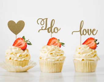May include: Three cupcakes with white frosting and a strawberry on top. Each cupcake has a gold glitter topper that says "I do" and "love". The cupcake on the left has a gold glitter heart topper.