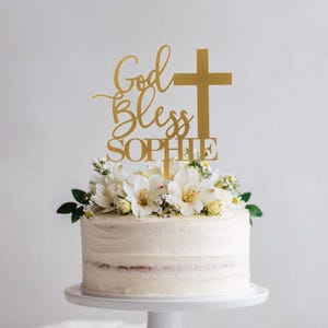 Pu&ograve; includere: Un topper per torta color oro con la scritta God Bless e il nome Sophie, accompagnato da un simbolo a croce. Il topper &egrave; posizionato su una torta con glassa bianca decorata con fiori bianchi, appoggiata su un'alzata per torte bianca.