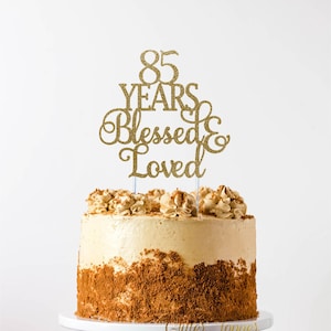 May include: Gold glitter cake topper with the text "85 Years Blessed & Loved". The topper is on top of a frosted cake with a brown crumb crust.