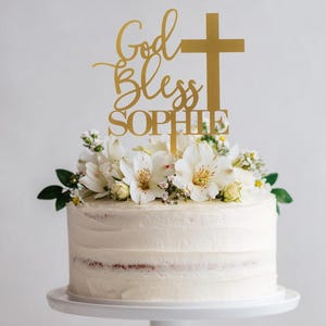 Pu&ograve; includere: Un topper per torta color oro con la scritta God Bless SOPHIE e il simbolo di una croce, posizionato sopra una torta con glassa bianca decorata con fiori bianchi. Il topper &egrave; realizzato in un materiale sottile e piatto, inserito al centro.