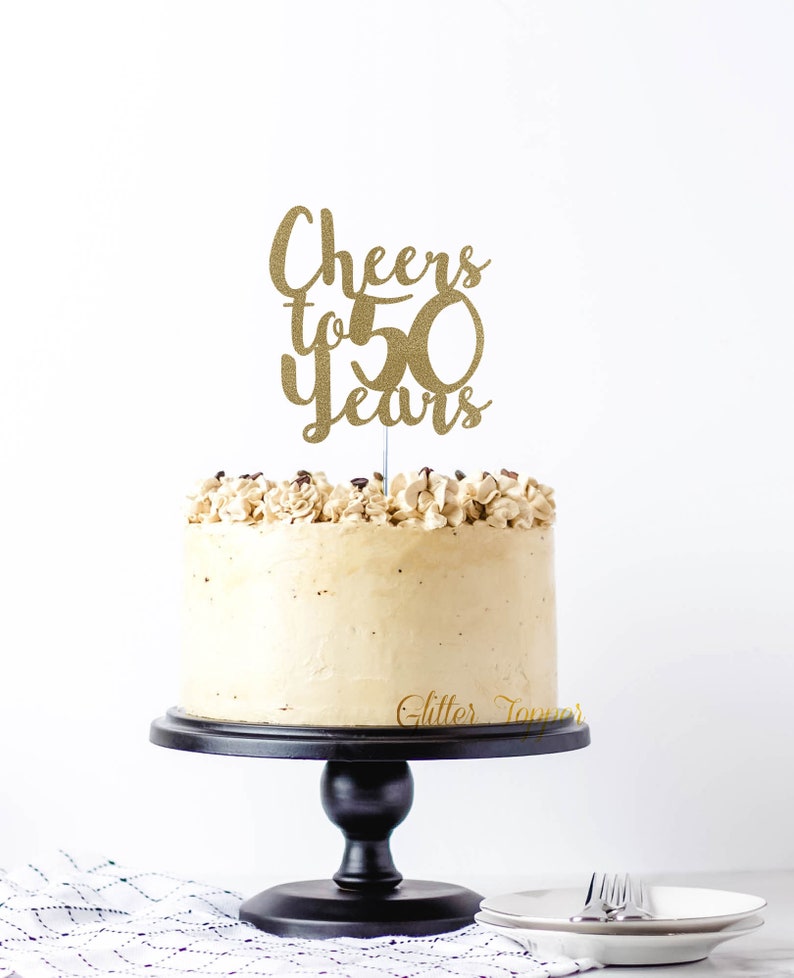 May include: Gold glitter cake topper with the text "Cheers to 50 Years" on a white cake with brown frosting.