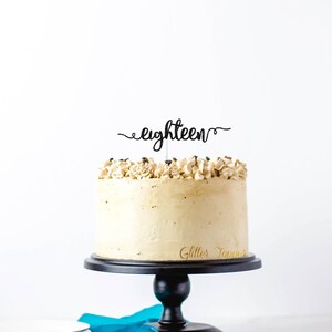 May include: A frosted cake with a black "eighteen" cake topper. The cake is on a black cake stand, with a blue cloth and silverware in the foreground. The cake has a light brown frosting and decorative frosting on top.