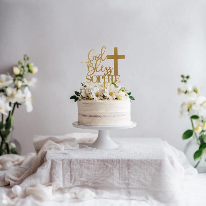 Pu&ograve; includere: Un topper per torta color oro con la scritta God Bless e il nome Sophie, accompagnato da un simbolo a croce. Il topper &egrave; posizionato su una torta con glassa bianca decorata con fiori bianchi, appoggiata su un'alzata per torte bianca.