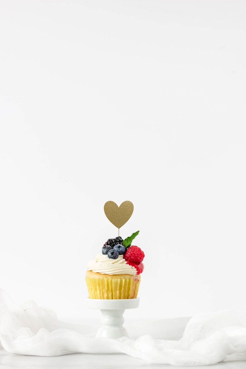 Op de afbeelding: Een enkele vanille cupcake bedekt met witte frosting en verse bessen, waaronder frambozen en blauwe bessen. Een gouden glitterhart zit bovenop de cupcake, die op een witte taartstandaard staat.